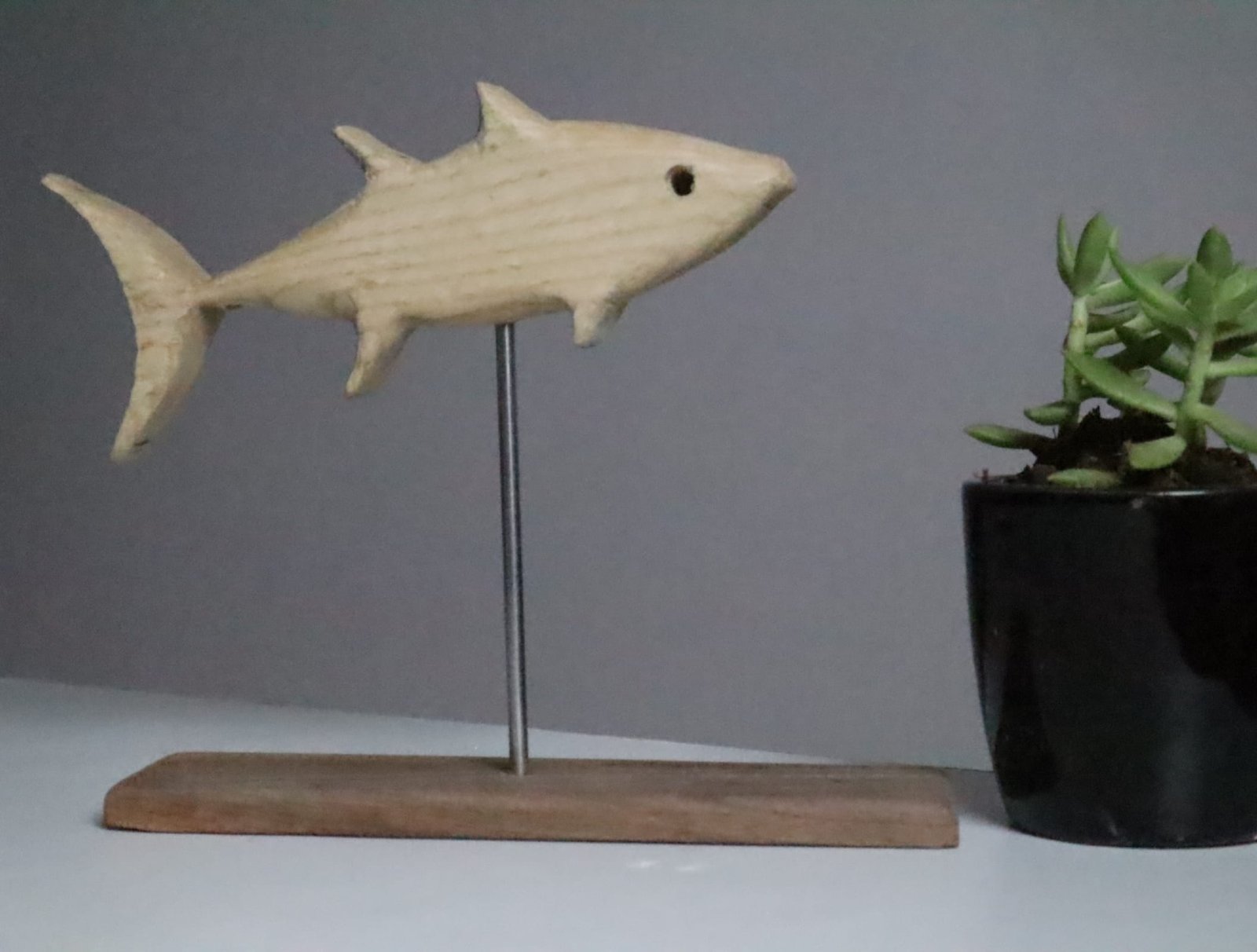 IMG_2286 Minimalist white ash wood fish sculpture mounted on stainless steel rod with wooden base, accompanied by a small succulent pot in a modern decor setting. Hand-carved eco-friendly wooden fish figurine displayed on tabletop with indoor plant, ideal for rustic and nature-themed interior styling. Artisan-crafted fish sculpture made of premium white ash wood, designed with clean lines and earthy accents for shelf or desk decor.