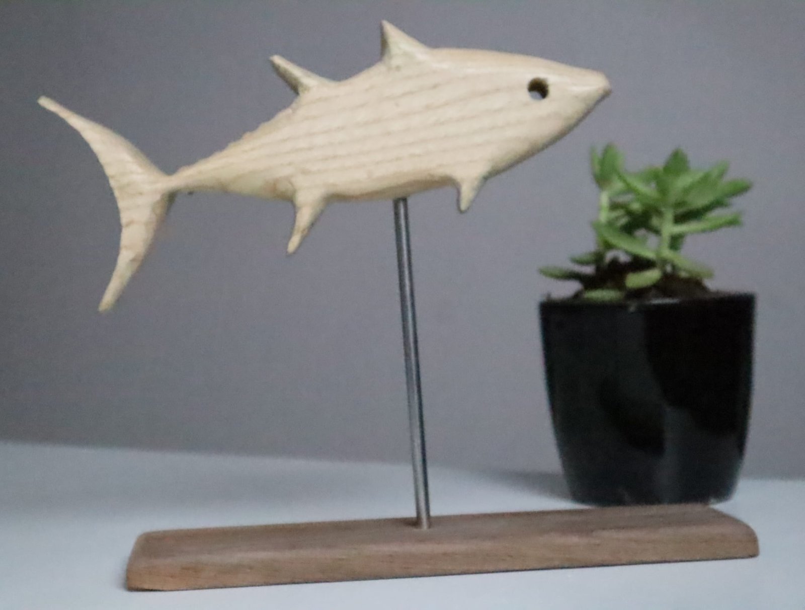 IMG_2287 Minimalist white ash wood fish sculpture mounted on stainless steel rod with wooden base, accompanied by a small succulent pot in a modern decor setting. Hand-carved eco-friendly wooden fish figurine displayed on tabletop with indoor plant, ideal for rustic and nature-themed interior styling. Artisan-crafted fish sculpture made of premium white ash wood, designed with clean lines and earthy accents for shelf or desk decor.